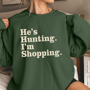 May include: A dark green sweatshirt with the text "He's Hunting. I'm Shopping." in a white, bold font. The sweatshirt has long sleeves and a relaxed fit, suitable for casual wear.