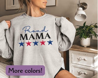 May include: A light gray sweatshirt with the words "Proud MAMA" in blue and black, and a row of red and blue stars. The image also includes a purple rectangle with the text "More colors!"