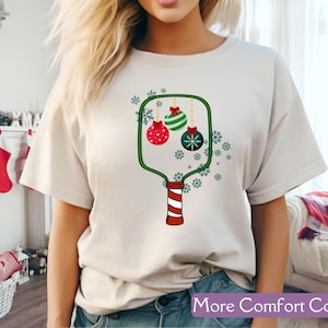 Puede incluir: Camiseta color crema con un dise&ntilde;o de adorno navide&ntilde;o. El gr&aacute;fico incluye adornos rojos, verdes y blancos, copos de nieve y el texto "More Comfort Colors" en una pancarta morada. El dise&ntilde;o est&aacute; impreso en una camiseta suave e informal.