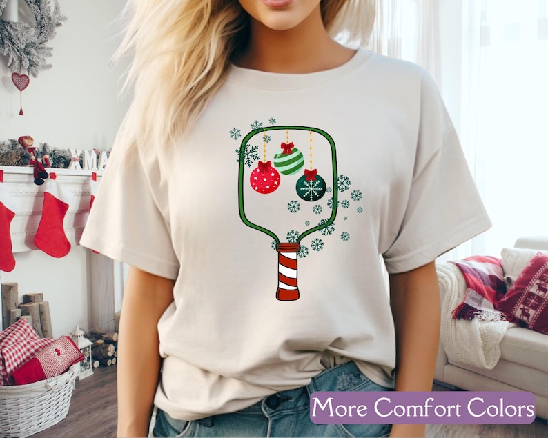 Puede incluir: Camiseta color crema con un dise&ntilde;o de adorno navide&ntilde;o. El gr&aacute;fico incluye adornos rojos, verdes y blancos, copos de nieve y el texto "More Comfort Colors" en una pancarta morada. El dise&ntilde;o est&aacute; impreso en una camiseta suave e informal.