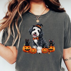 May include: Dark gray t-shirt featuring a Halloween-themed graphic. The design includes a black and white dog wearing a witch hat and orange bow tie, surrounded by jack-o'-lanterns and black cats. The shirt is made of a soft, comfortable material.