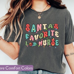 May include: Dark gray t-shirt with the text "SANTA'S FAVORITE L&D NURSE" in colorful, retro-style lettering. The design includes string lights. The shirt is a comfort color style.
