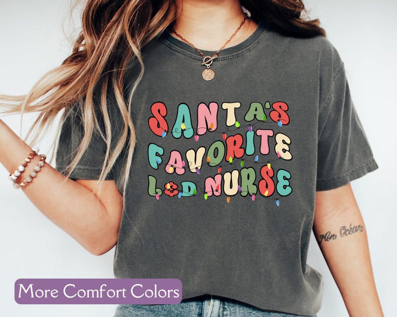 May include: Dark gray t-shirt with the text "SANTA'S FAVORITE L&D NURSE" in colorful, retro-style lettering. The design includes string lights. The shirt is a comfort color style.
