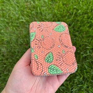 May include: A small, rectangular, peach-colored box with a repeating pattern of green leaves, orange suns, and white stars. The box is made of a textured material and has a shiny finish.