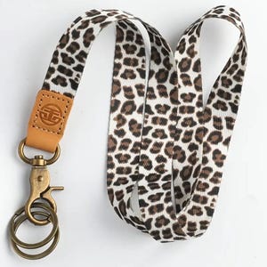 May include: A lanyard featuring a leopard print pattern in brown and black on a white background. It has a tan leather accent with a logo, a gold-coloured clasp, and two key rings. The lanyard is designed to be worn around the neck.