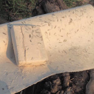 Puede incluir: Una pastilla de jabón rectangular blanquecina y una hoja de papel a juego, ambas con un estampado de hojas. El jabón descansa sobre el papel, que está ligeramente curvado. Los artículos están sobre una superficie de madera natural.