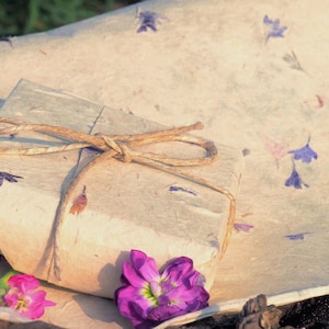 Op de afbeelding: Een ingepakt cadeau vastgebonden met touw, liggend op papier met verspreide bloemaccenten. Het cadeau is verpakt in getextureerd papier met een natuurlijke uitstraling. Heldere roze en paarse bloemen staan op de voorgrond.
