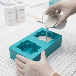 May include: A teal silicone mould with two cavities is being filled with a light grey liquid from a glass beaker. Two white bottles labelled "PART A" and "PART B" are visible in the background, along with other tools.