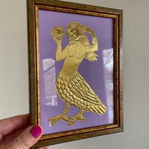 May include: A gold-colored framed print of a mermaid holding a round object. The mermaid is depicted with a human torso and a bird-like lower body with feathers. The background is a light purple color.