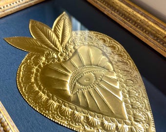 May include: A gold-colored heart-shaped metal wall hanging with an eye design in the center. The heart is framed in a gold-colored frame with a dark blue background.