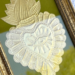 Puede incluir: Un corazón de metal dorado con un diseño de ojo y rayos de sol, enmarcado en un marco dorado sobre un fondo verde.