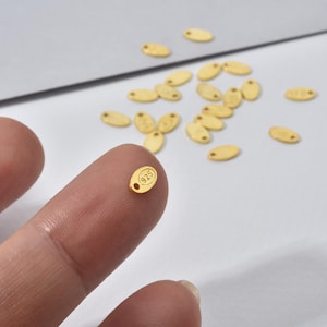 May include: Close-up of small, gold-colored oval tags. One tag is held between fingers, displaying the number "925" in a circle. The tags have a small hole at one end. Several other tags are scattered on a light-colored surface.
