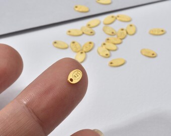 May include: Close-up of small, gold-colored oval tags. One tag is held between fingers, displaying the number "925" in a circle. The tags have a small hole at one end. Several other tags are scattered on a light-colored surface.