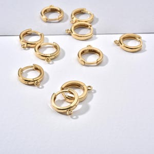 May include: Several gold-coloured hoop earrings, each with a small loop for attaching charms. The earrings are displayed on a white surface, highlighting their simple design. The earrings are small and round, suitable for everyday wear.