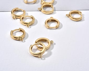 以下が含まれることがあります： 小さなチャームなどを取り付けるための小さなループが付いた、金色のフープピアスです。シンプルなデザインが際立つように、白い表面に配置されています。ピアスは小さく丸みを帯びています。
