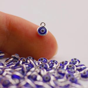 May include: A collection of small, round charms with a blue and white eye design. Each charm has a silver-colored metal border and a small loop at the top. The charms are scattered on a white surface.