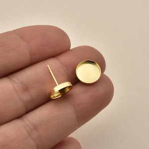 Könnte beinhalten: Zwei goldfarbene Ohrstecker-Komponenten. Ein Ohrring hat einen Stift zum Einsetzen, der andere ist eine flache, kreisförmige Fassung. Die Ohrringe sind so konzipiert, dass sie einen Stein oder ein anderes dekoratives Element halten. Sie sind vor neutralem Hintergrund.