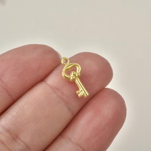 May include: A small gold-colored metal key charm featuring a decorative bow with three small spheres at the base of the head and a simple bit at the bottom. The charm includes a small circular loop at the top for attachment to jewelry or crafts.