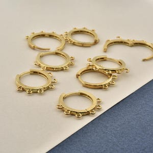 Può includere: Diversi orecchini a cerchio dorati con piccoli dettagli decorativi sono sparsi su una superficie. Gli orecchini presentano un design circolare semplice con molteplici abbellimenti arrotondati.