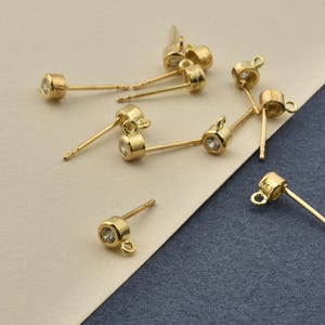 以下が含まれることがあります： ゴールドトーンのスタッドピアス用パーツのコレクション。それぞれベゼルにセットされた小さな透明な円形の石が特徴です。各スタッドにはポストとベース部分に小さなループが付いており、他のジュエリーパーツを取り付けることができるようになっています。