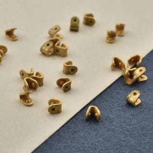 May include: A collection of small, U-shaped brass metal crimp ends with a matte finish. Each piece features a curved design with a single circular hole at the top, intended for securing cord or thread ends in jewelry making and craft projects.