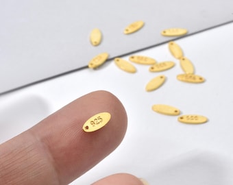 May include: Close-up of small, oval-shaped gold-colored metal tags. Each tag has the number "925" stamped on it, indicating the metal's purity. The tags are scattered, with one held between fingers. These are likely jewelry components.