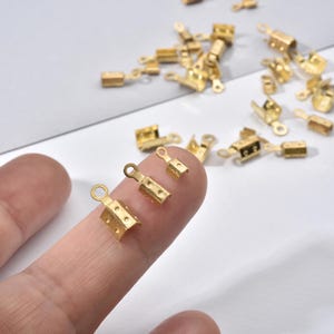 May include: A collection of small, gold-colored crimp end findings for jewelry making. The findings have a rectangular shape with holes for attaching to cords or chains. Some are held on a finger.