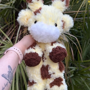 May include: A soft, plush giraffe toy in cream and brown. The giraffe has brown spots, hooves, and ears, with a white muzzle and black eyes. The toy is designed to be cuddly and is made with a textured material.