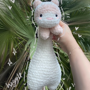 May include: A handmade plush toy in the shape of a bear, featuring a white body and a pink and white head. The toy has small black eyes and a stitched smile. The background is a blurred view of green foliage. Text on the image says "Wiggle!"