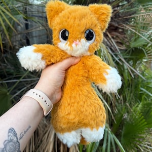 May include: A plush cat toy with orange fur, large black eyes, a small pink nose, and white patches on the face, paws, and lower body. The stuffed animal has a soft, fuzzy texture and pointed ears, designed with a rounded head and slender body.