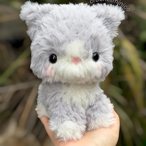 May include: A plush cat toy with soft, shaggy grey and white fur, featuring large black plastic eyes, a small pink nose, and light pink blush on the cheeks. The text crochetminico is visible in the upper right corner of the image.