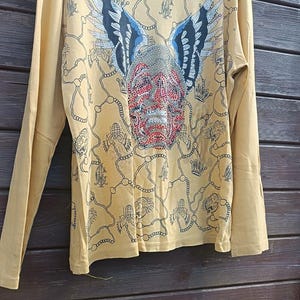May include: Mustard yellow long-sleeve shirt with a detailed graphic on the front. The graphic features a face with red, silver, and black accents, wings, and chain-like patterns. The shirt is hanging on a pink hanger.