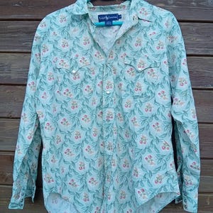 Può includere: Camicia a maniche lunghe con motivo floreale nei toni dell'azzurro, rosa e verde. La camicia ha due tasche sul petto ed è appesa a un'appendiabiti blu. L'etichetta del marchio è visibile.