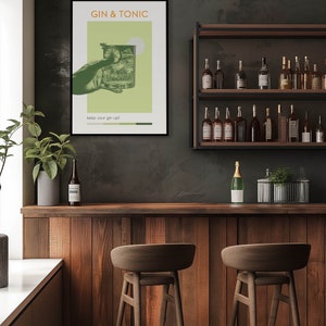 The green color Gin Tonic print, carries retro vibe effects, a hand holding a cocktail glass, hanging on a wall on the counter in front of the modern minimalist bar. There are cocktail bottles, plants, and brown bar stools on the side