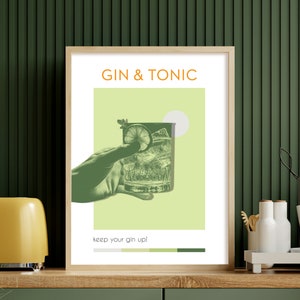 The minimalist green color Gin Tonic print, carries modern vibe effects, a hand holding a glass, leaning against the green wall on the counter of a modern kitchen. There are espresso cups and a toaster on the side.