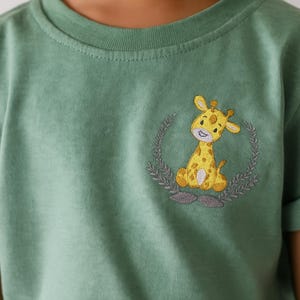 May include: A sage green t-shirt featuring an embroidered yellow giraffe design. The giraffe is inside a gray wreath. The shirt has short sleeves and a crew neck.