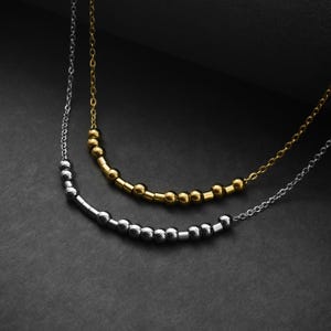 May include: Two delicate necklaces featuring thin metal chains. One necklace is gold-toned with a series of small round and cylindrical beads arranged in a pattern. The second necklace is silver-toned with a similar arrangement of beads.