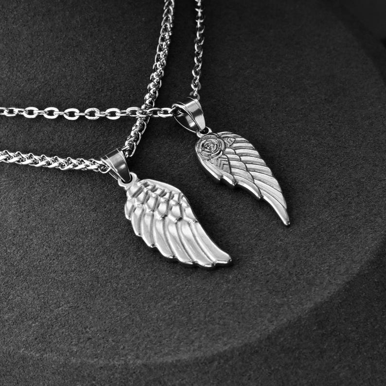 May include: Two silver-colored necklaces with matching wing pendants. Each pendant features detailed feathering and is suspended from a delicate chain. The necklaces are displayed against a dark background.
