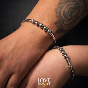 May include: Two silver bracelets with a beaded design are shown on wrists. The bracelets have a series of small, round beads and cylindrical links. The bracelets are on a dark background. The text "LOVE BRASLET" is at the bottom.