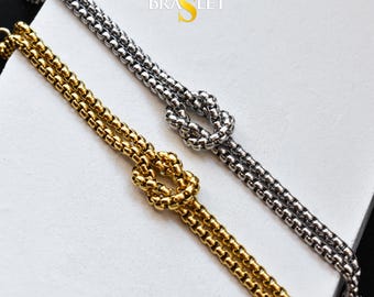 Peut inclure: Gros plan sur deux bracelets, un doré et un argenté, chacun avec un motif de nœud. Les bracelets sont faits de petits maillons métalliques connectés. Le mot "LOVE BRASLET" est imprimé en haut à droite.