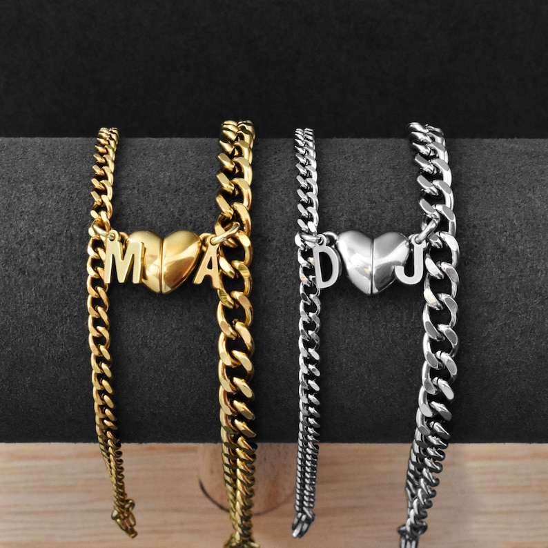 May include: Two curb chain bracelets, one gold-toned and one silver-toned, each featuring a central heart-shaped charm. The gold bracelet displays the letters M and A on the heart, while the silver bracelet displays the letters D and J on the heart.