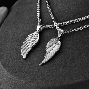May include: Two silver-toned wing pendant necklaces. One wing pendant is plain, while the other features a rose design. The necklaces are displayed on a dark gray surface, with the chains visible. The pendants are detailed and reflective.