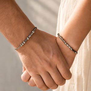 Può includere: Due braccialetti abbinati caratterizzati da una serie di perline sferiche color argento lucido alternate a piccoli distanziatori cilindrici. I braccialetti sono realizzati con un delicato design a maglie di catena e un fermaglio di chiusura.