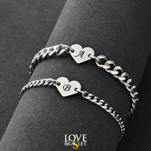 Peut inclure: Deux bracelets en chaîne argentés avec des pendentifs en forme de cœur. L'un porte la lettre "A", l'autre "B". Les bracelets sont présentés sur une surface gris foncé. La marque "LOVE BRASLET" est en bas.