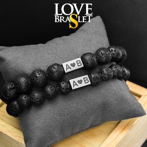 Op de afbeelding: Twee zwarte lavasteen kralenarmbanden met zilveren rechthoekige bedels, elk gegraveerd met "A♥B". De armbanden liggen op een grijs kussen. De tekst "LOVE BRASLET" staat bovenaan de afbeelding.