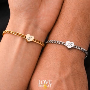 May include: Two matching bracelets, one gold-toned and one silver-toned, each featuring a heart-shaped charm with an initial. The gold bracelet has the letter "G", and the silver bracelet has the letter "D". Both bracelets have a curb chain design.