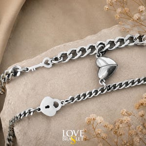 Könnte beinhalten: Zwei silberfarbene Metallkettenarmbänder mit einem herzförmigen Anhänger, der sich in zwei Teile teilen lässt. Ein Armband hat einen kleinen Schlüsselanhänger, das andere ein herzförmiges Vorhängeschloss. Der Text LOVE BRASLET ist sichtbar.