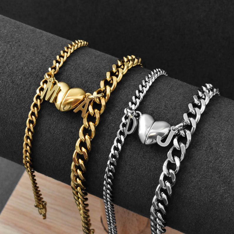 May include: Two curb chain bracelets, one gold-toned and one silver-toned, each featuring a split heart charm. The gold bracelet displays the letters M and A, while the silver bracelet displays the letters D and J attached to the heart halves.
