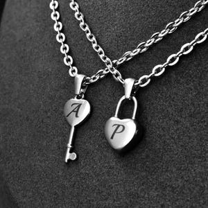 May include: Two silver necklaces with heart-shaped pendants. One pendant is a key with the letter "A", and the other is a lock with the letter "P". The necklaces hang on a silver chain.