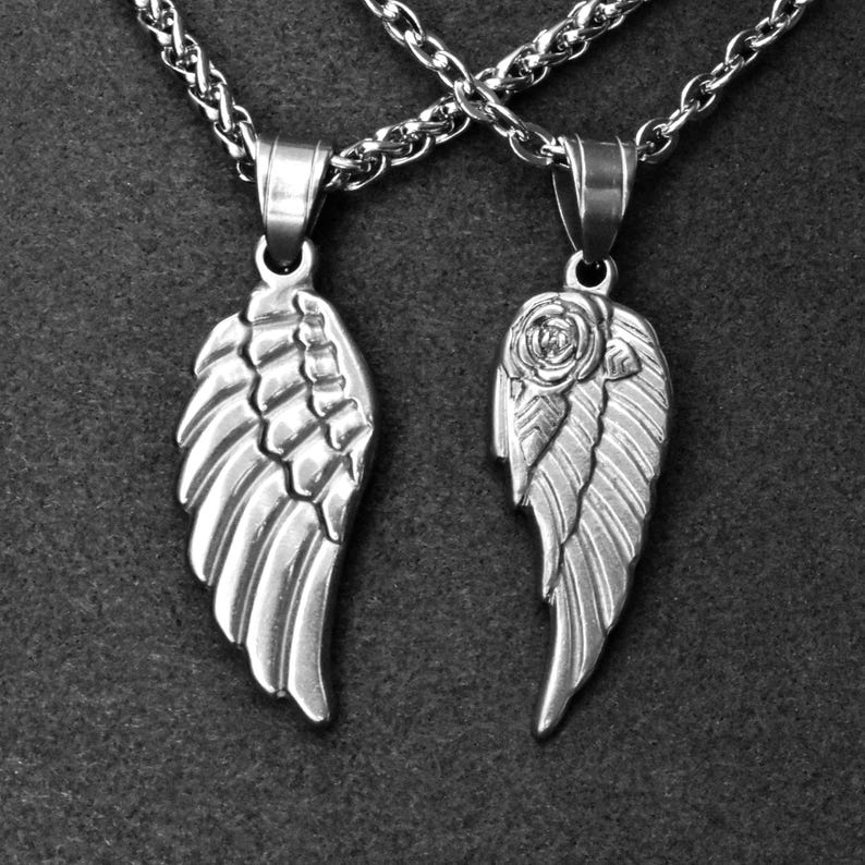 May include: Two silver-toned wing pendant necklaces. One wing features detailed feathering, the other has a rose at the top. Both hang from a silver chain. The pendants are displayed against a dark gray background.